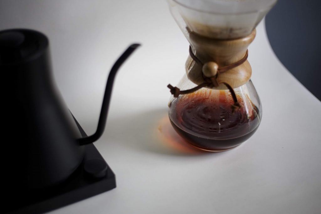 Freshly brewed hot coffee inside a Chemex coffeemaker alongside a black Fellow kettle